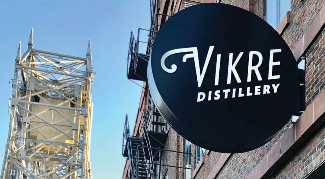 A sign with "Vikre Distillery" on it, in front of a steel bridge structure, against a clear sky