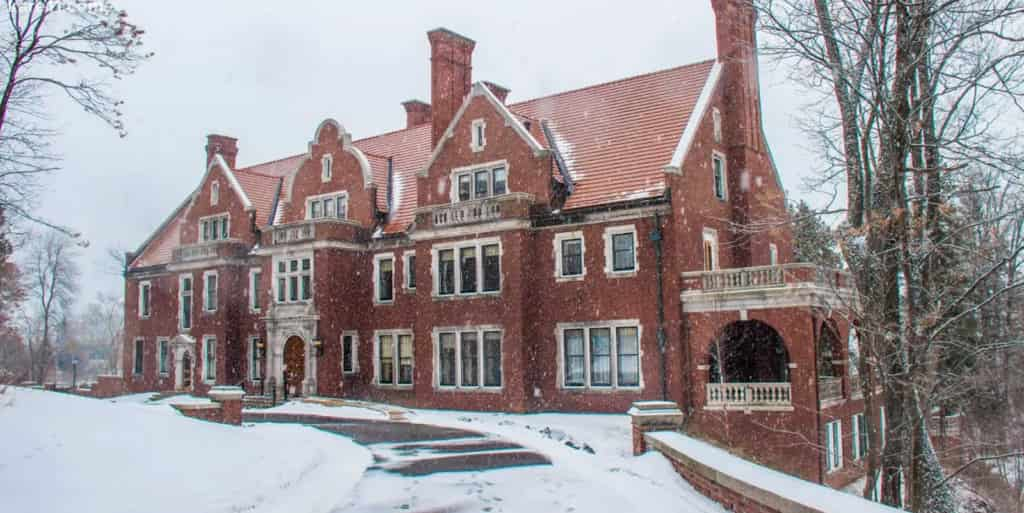 Snow gently falls on a historic red-brick mansion with multiple chimneys