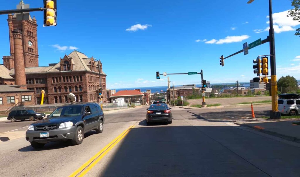Traffic flows through an intersection in Duluth with a historic building and lake view