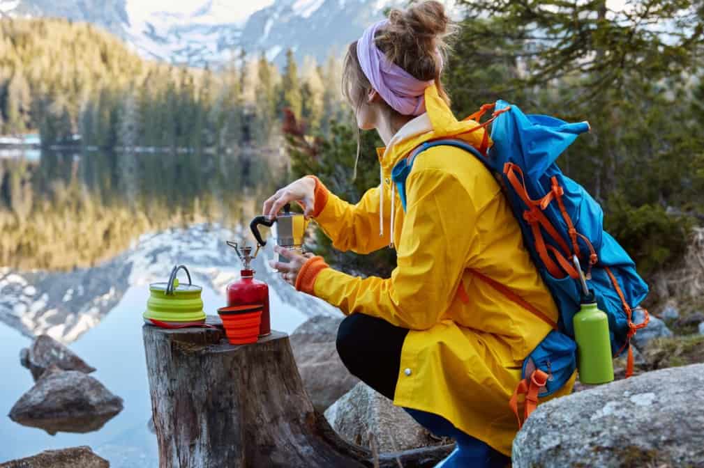 A hiker prepares a drink on a camp stove by a mountain lake
